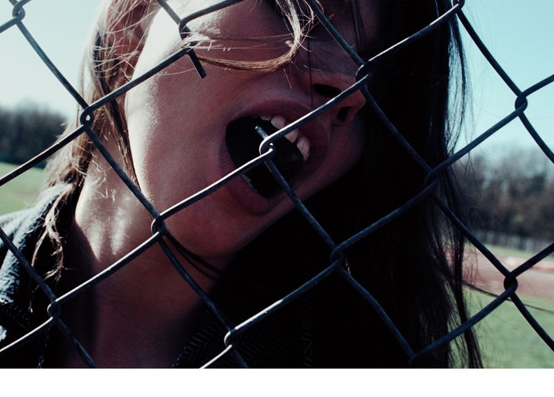 Teenage girl with mouth open and teeth visible stands behind a chain link fence outdoors on a sunny day; the background is blurry.