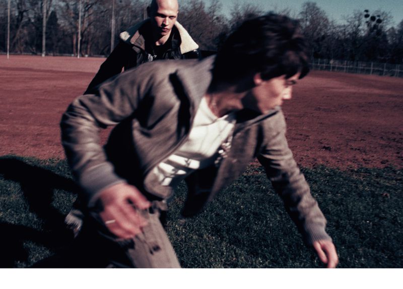Two men in a field: one running toward the viewer in a brown jacket, the other behind him, bald, in a darker jacket.