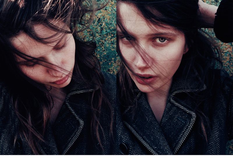 A portrait photograph shows a woman with fair skin and long, loose brown hair wearing a dark grey jacket, laying on a textured rug.