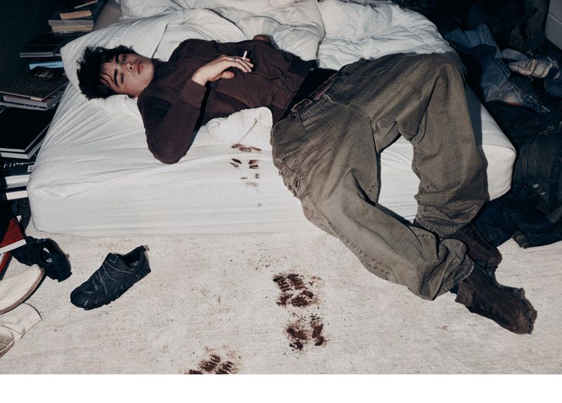 Adult male lying on a white bed, holding a cigarette, wearing brown shirt and jeans; dark marks on bed and floor, messy room. 