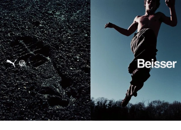 Athletic barefoot man in mid-air, set against a bright blue sky, with a foreground of dark gravel and sports brand logos.