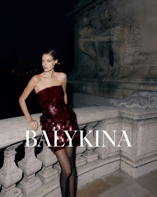 Elegant woman in a red strapless sequin dress and black stockings poses against a stone structure at night, showcasing fashion and style.