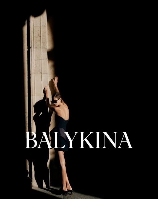 A fashionable woman poses in a black mini dress, sunglasses, and heels next to a textured column in a dark, contrasted environment.