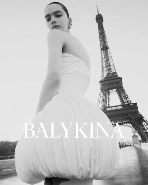 Woman poses in a white dress with the Eiffel Tower in the background. The black and white photograph highlights the fashion and architecture.