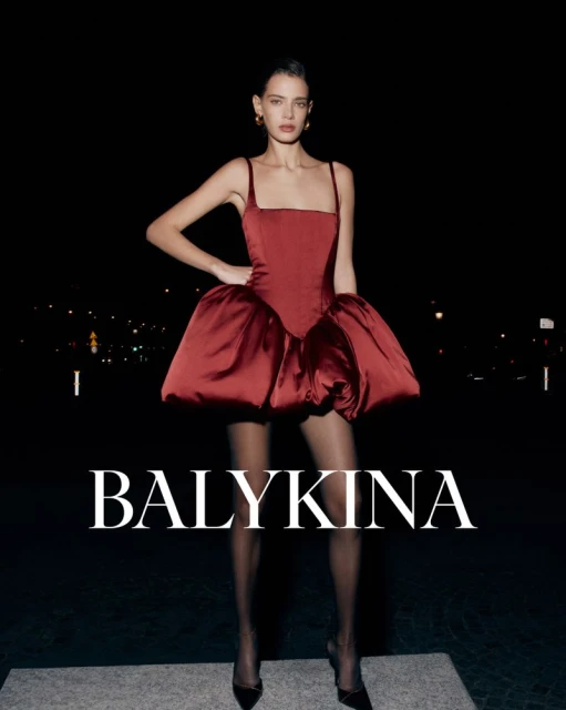A stylish woman in a red satin mini dress and heels stands on a concrete surface against a dark night cityscape backdrop.
