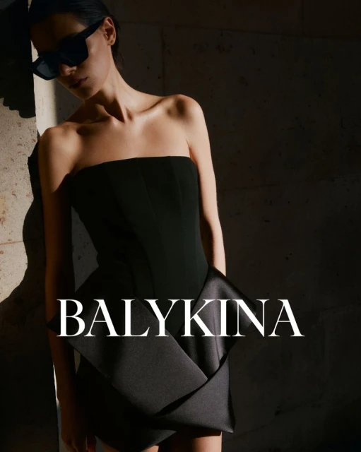 Woman in strapless black dress and sunglasses leans against a stone wall, showcasing the Balykina brand logo in a fashion editorial shot.