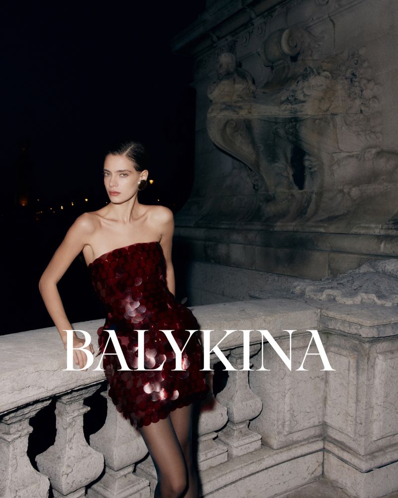 A woman in a strapless maroon sequined dress and dark stockings poses against an ornate stone wall with the logo 'BALYKINA'.
