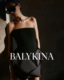 Model wearing a black strapless dress and dark sunglasses leans against a stone wall with dramatic lighting.