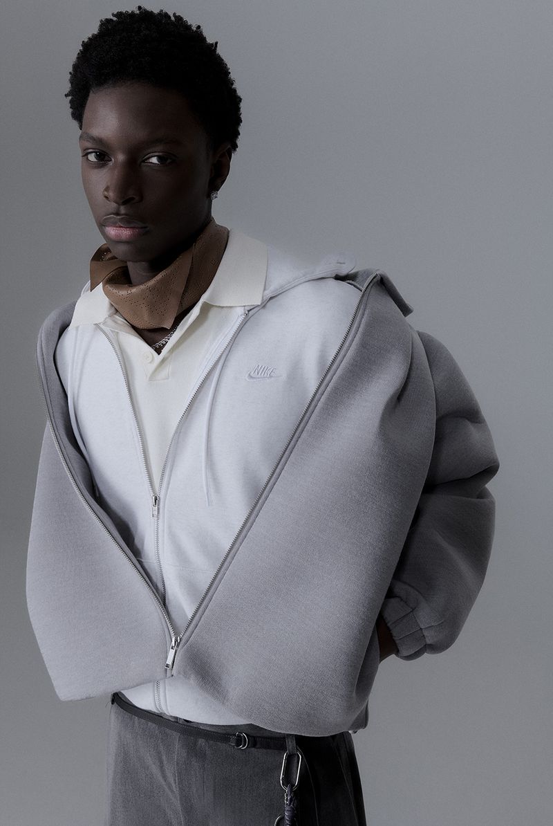 A young Black male model poses in an indoor studio with grey background, wearing an open grey zip-up hoodie, collared shirt, and grey skirt.