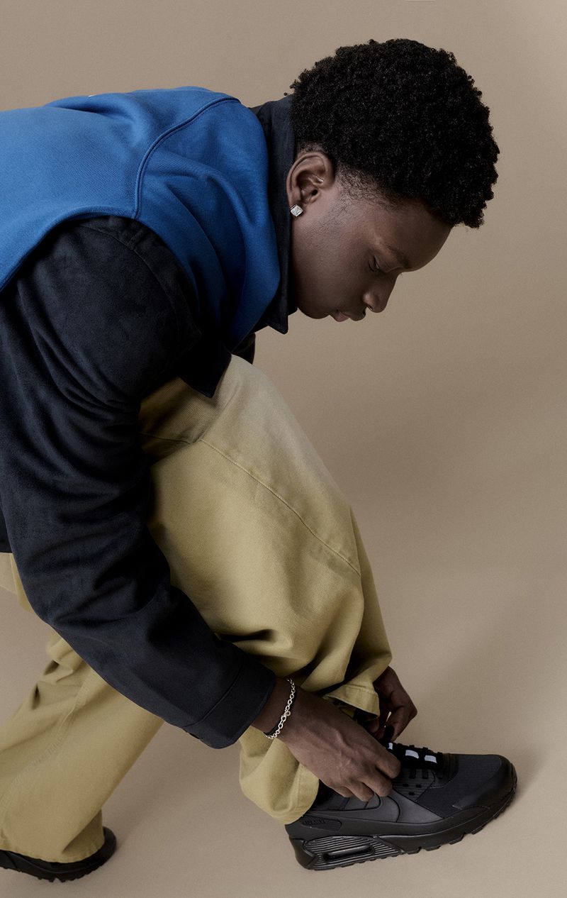 An African-American man with dark curly hair is tying the laces of a black Nike sneaker, wearing baggy yellow pants and a blue jacket.