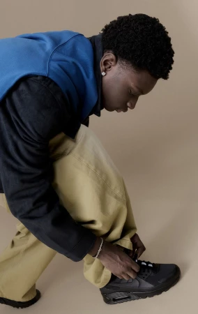 An African-American man with dark curly hair is tying the laces of a black Nike sneaker, wearing baggy yellow pants and a blue jacket.