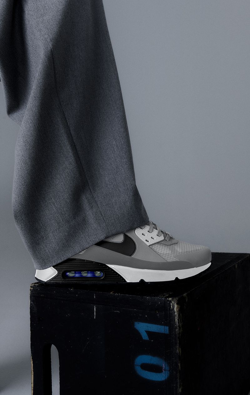 Grey Nike Air Max 90 sneaker on a black wooden box against a grey background, paired with grey trousers for a minimalist fashion shot.