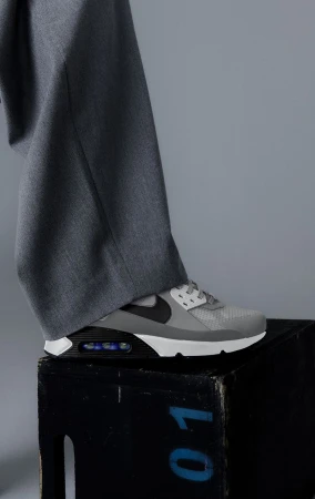 Grey Nike Air Max 90 sneaker on a black wooden box against a grey background, paired with grey trousers for a minimalist fashion shot.