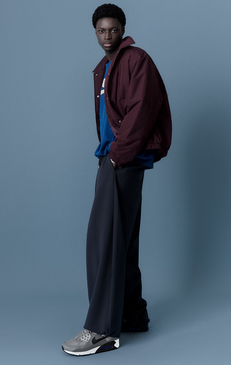 Male fashion model in a maroon jacket, blue shirt, and grey sneakers poses against a blue background in a studio shot.