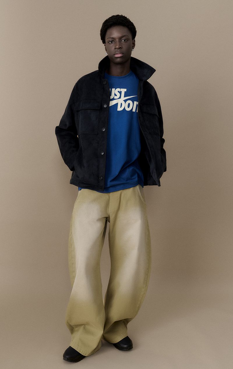 An African American man in a navy jacket, blue Nike tee, and tan wide-leg pants poses in a studio with a neutral background.
