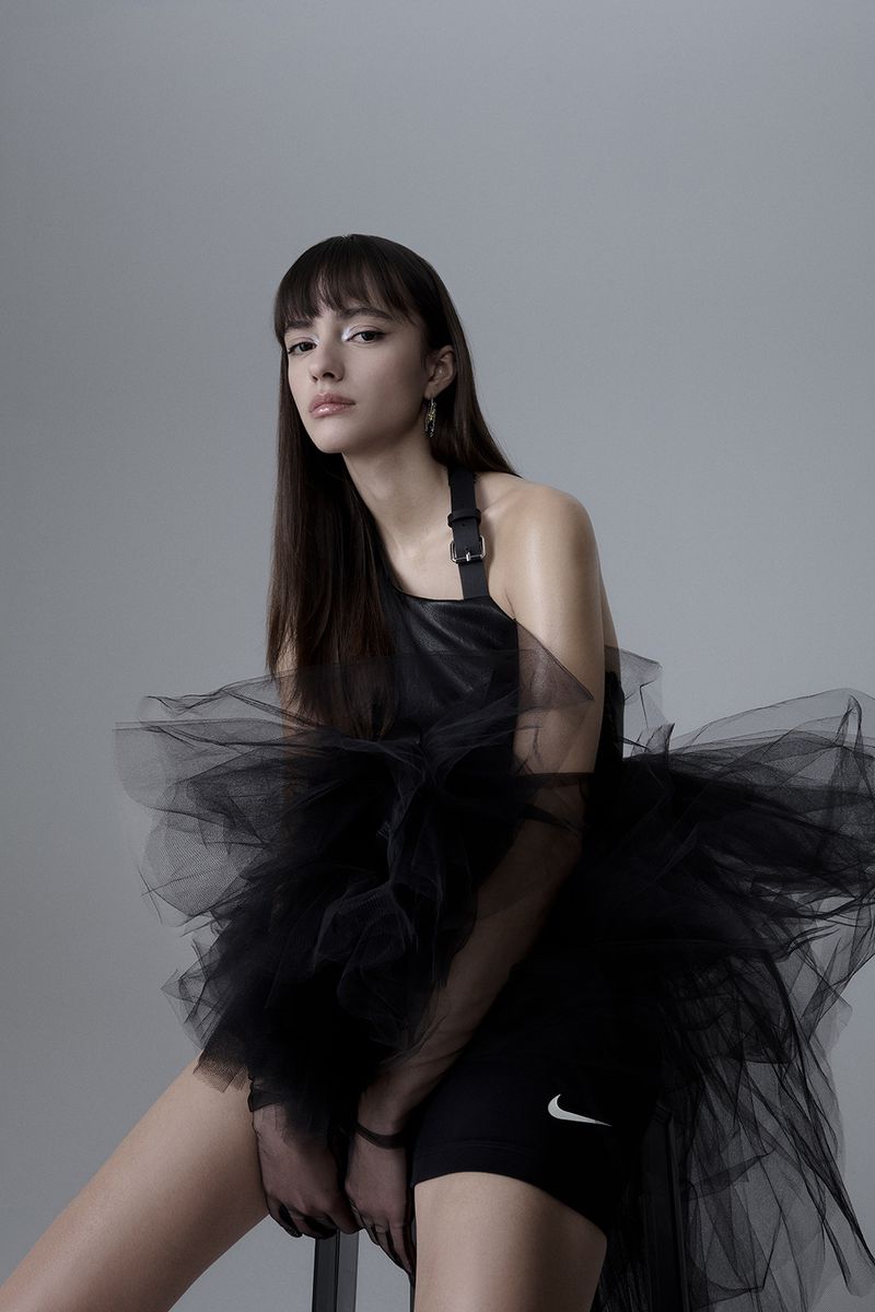 Portrait of a slender young woman with bangs and long brown hair wearing a black tulle dress and black Nike cycling shorts in studio.