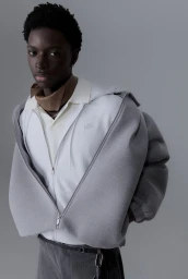 Black man wearing a grey zip hoodie over a white polo shirt against a plain grey background
