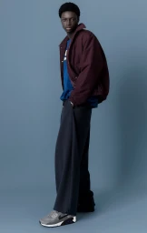 Young Black man standing against a blue background wearing a burgundy jacket over a blue sweater and grey sneakers