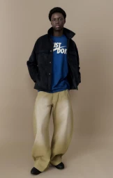 Black man standing against a neutral studio backdrop wearing a dark jacket, blue t-shirt, and pale trousers.