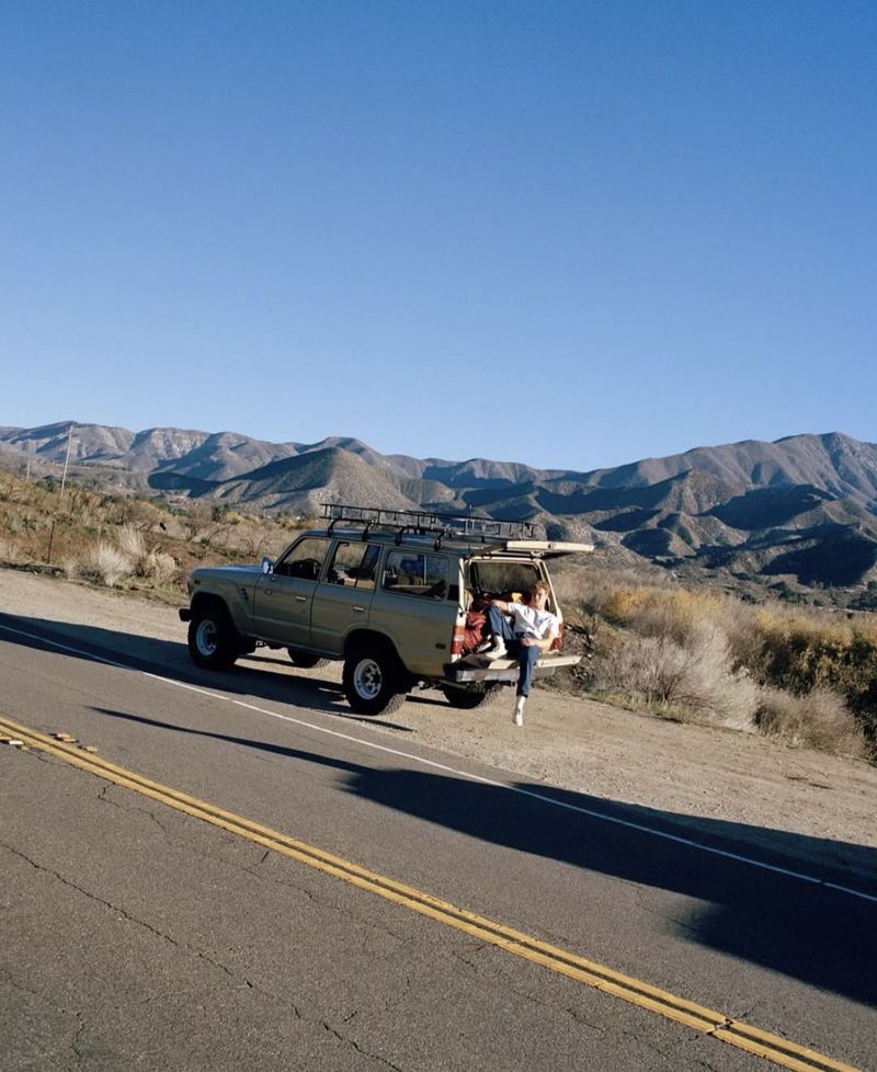 SUV parked on the side of a road with mountains in the distance. Woman sitting in the back with her legs hanging out.