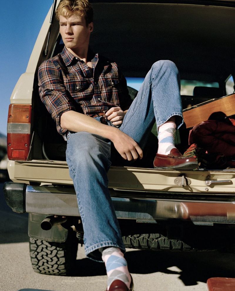Young adult male in a plaid shirt and jeans sits on the open tailgate of a tan SUV with blue sky in the background in this outdoor fashion shot.