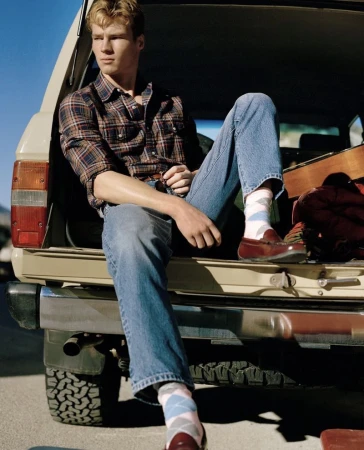 Young adult male in a plaid shirt and jeans sits on the open tailgate of a tan SUV with blue sky in the background in this outdoor fashion shot.