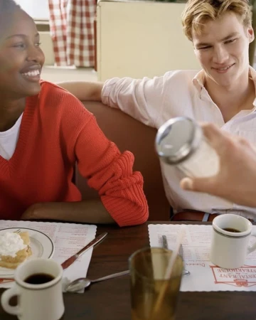 Two people smiling in a diner while a third person pours sugar into a glass. Retro diner scene with people, sugar, and coffee cups.