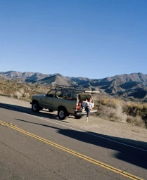 Model seated in the open tailgate of an SUV parked on a roadside with mountain range backdrop.