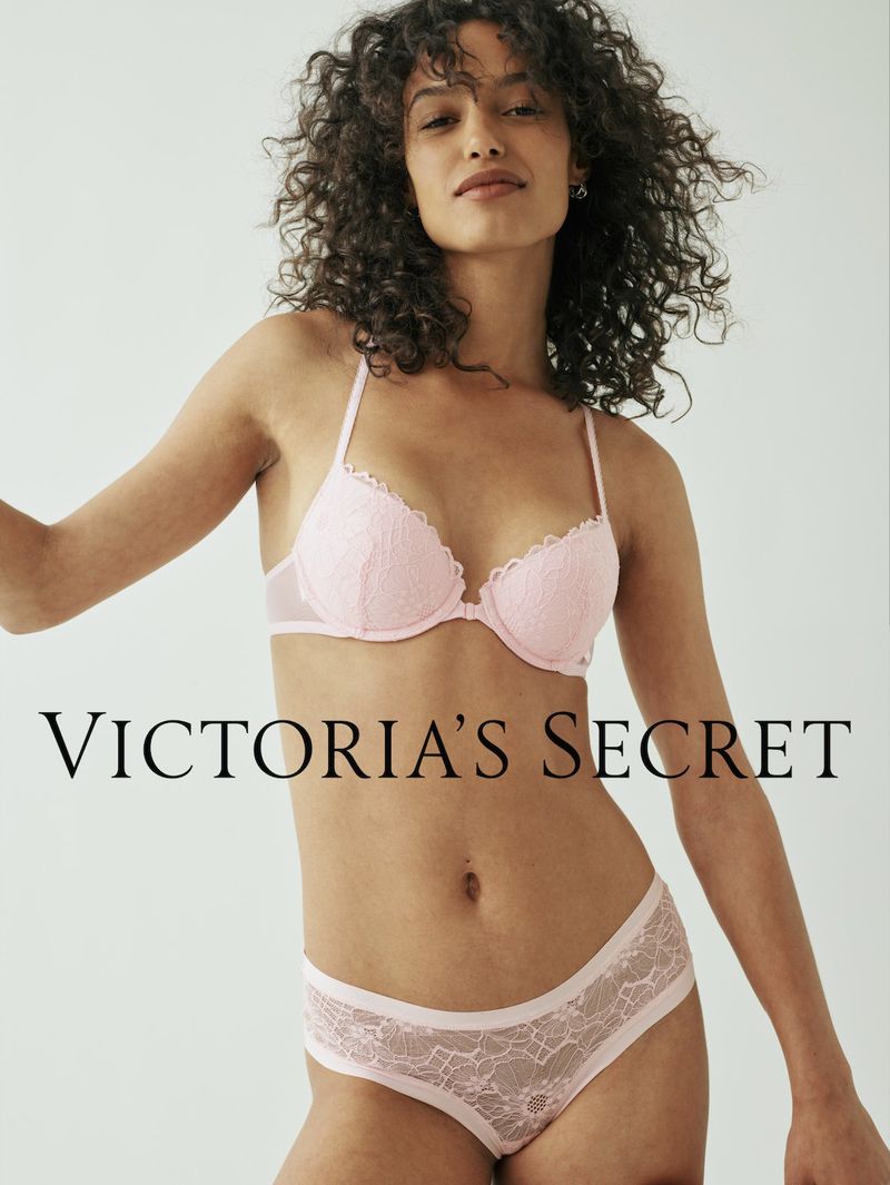 A woman poses in a pink lace bra and panty set, standing in front of a neutral background with the Victoria's Secret logo visible.