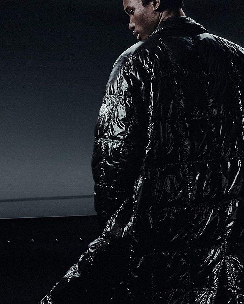 Fashion shot: A man wearing a shiny, black, puffy coat standing indoors against a dark, solid background in a studio setting.