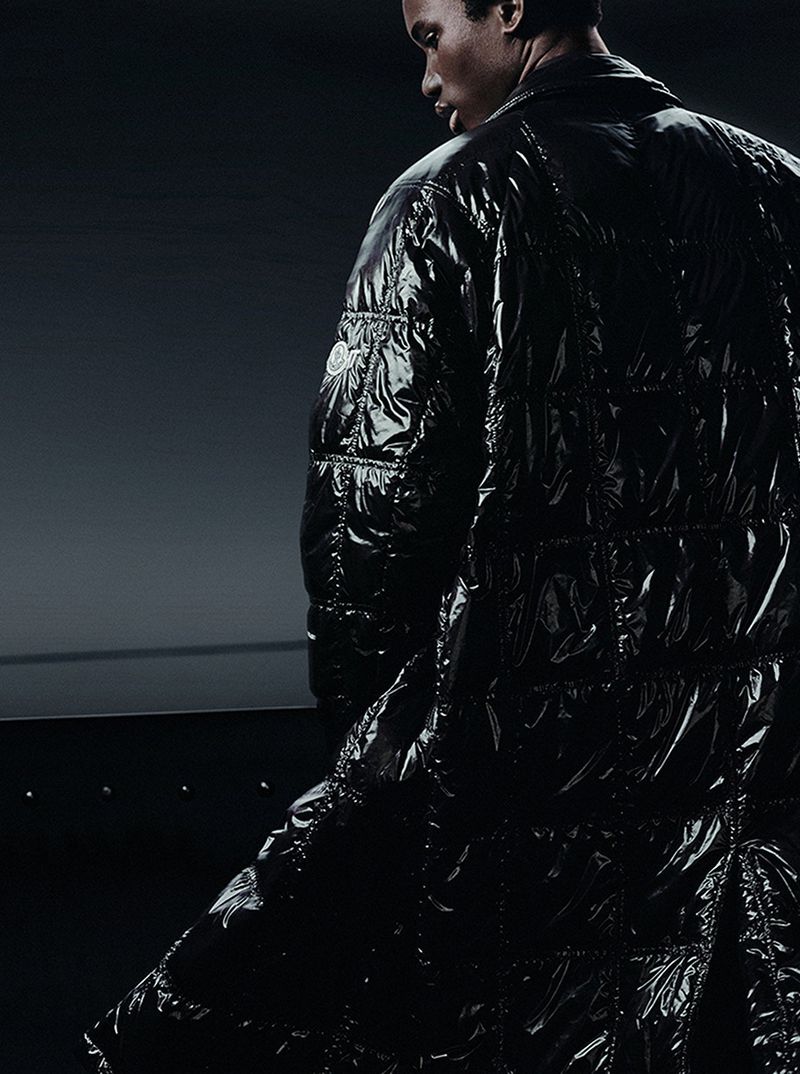 Male fashion model posing in a long, glossy black puffer coat against a dark background in a studio fashion editorial-style shot.
