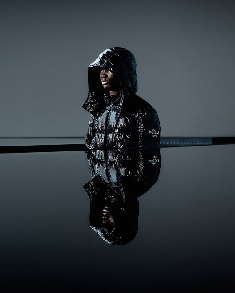 Studio shot of a dark-skinned male model in a glossy black hooded puffer jacket against a gray background, reflected in a dark surface.