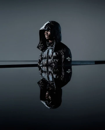 Studio shot of a dark-skinned male model in a glossy black hooded puffer jacket against a gray background, reflected in a dark surface.
