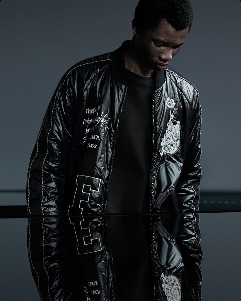 A dark-skinned male in a black bomber jacket with white writing and embroidery, standing indoors against a gray wall with reflective surface.