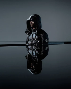 Model Alexander Acquah wearing a glossy black hooded puffer jacket with a sharp reflection in still water by photographer Leslie Zhang for Moncler Genius