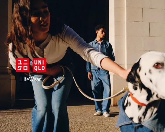 Woman walks Dalmatian wearing a denim jacket in sunny urban setting. Man in denim jacket stands behind. Fashion ad for Uniqlo.
