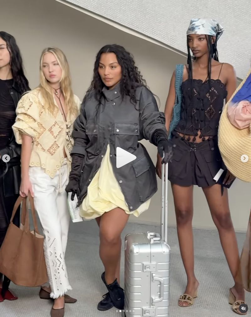 A group of diverse women in fashionable outfits pose with luggage against a modern tiled wall indoors during daytime, showing off style.