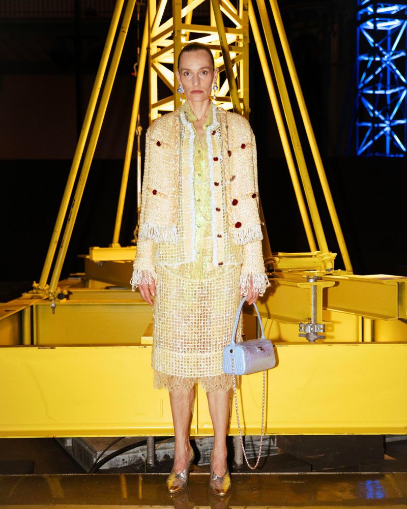 Model wearing tweed jacket and skirt standing before a large yellow industrial structure