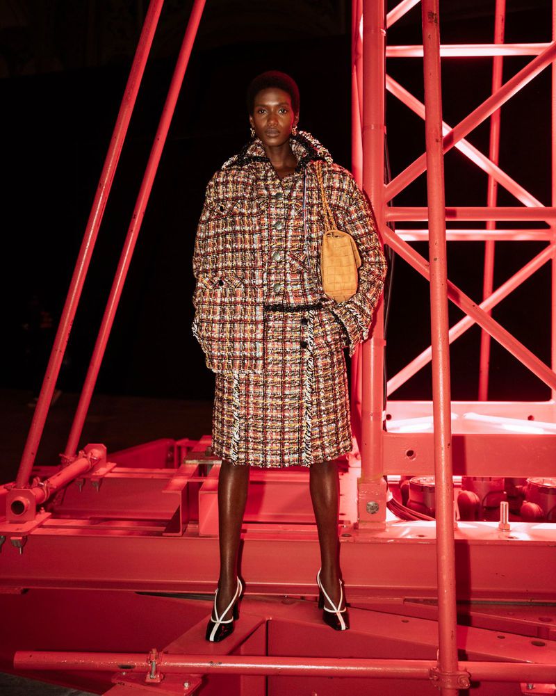 A dark-skinned fashion model poses in a tweed skirt suit with an amber purse against a red abstract background, creating a stark contrast.