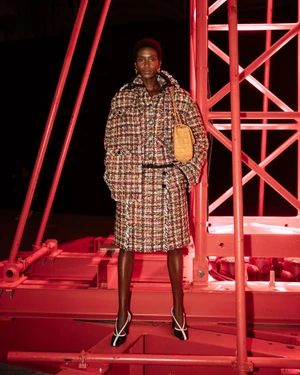 Model standing on a bright red industrial structure wearing a tweed jacket and matching skirt holding a tan handbag.