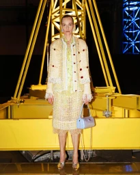 Model wearing tweed jacket and skirt standing before a large yellow industrial structure