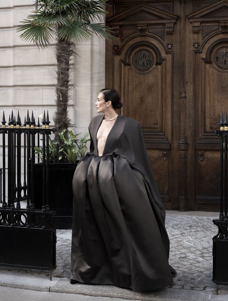 Elegant woman in a dark, flowing gown with a plunging neckline poses in front of an ornate wooden door with a palm tree nearby.