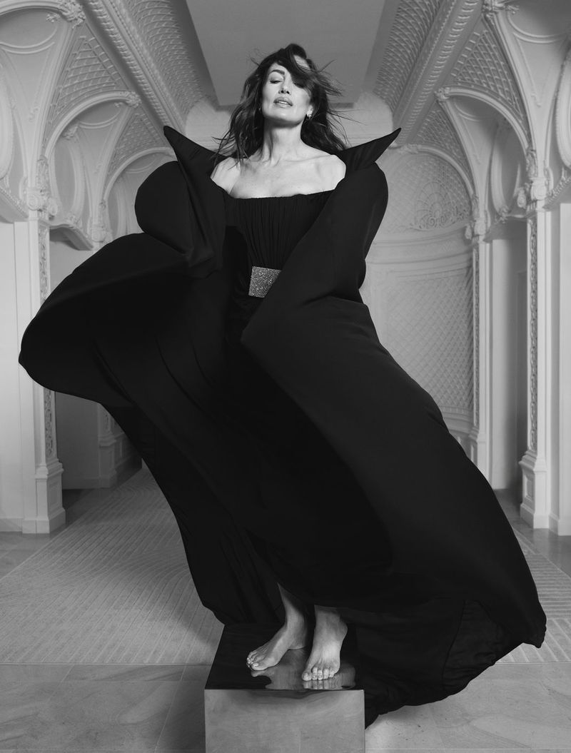 Monochromatic portrait of a woman in a black dress with dramatic sleeves, standing on a platform in an ornate interior space.