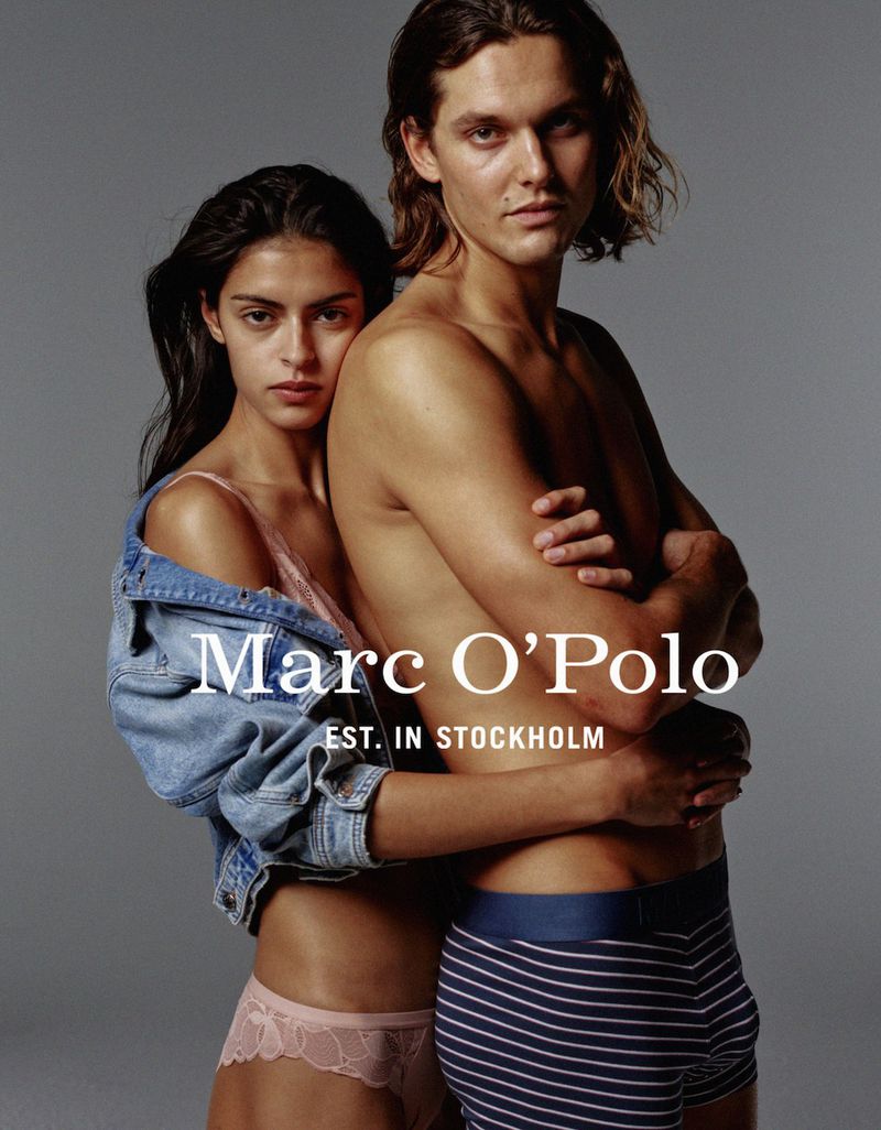Fashion shot of a couple embracing in underwear, woman in jean jacket. The couple is in a studio with a gray backdrop. 