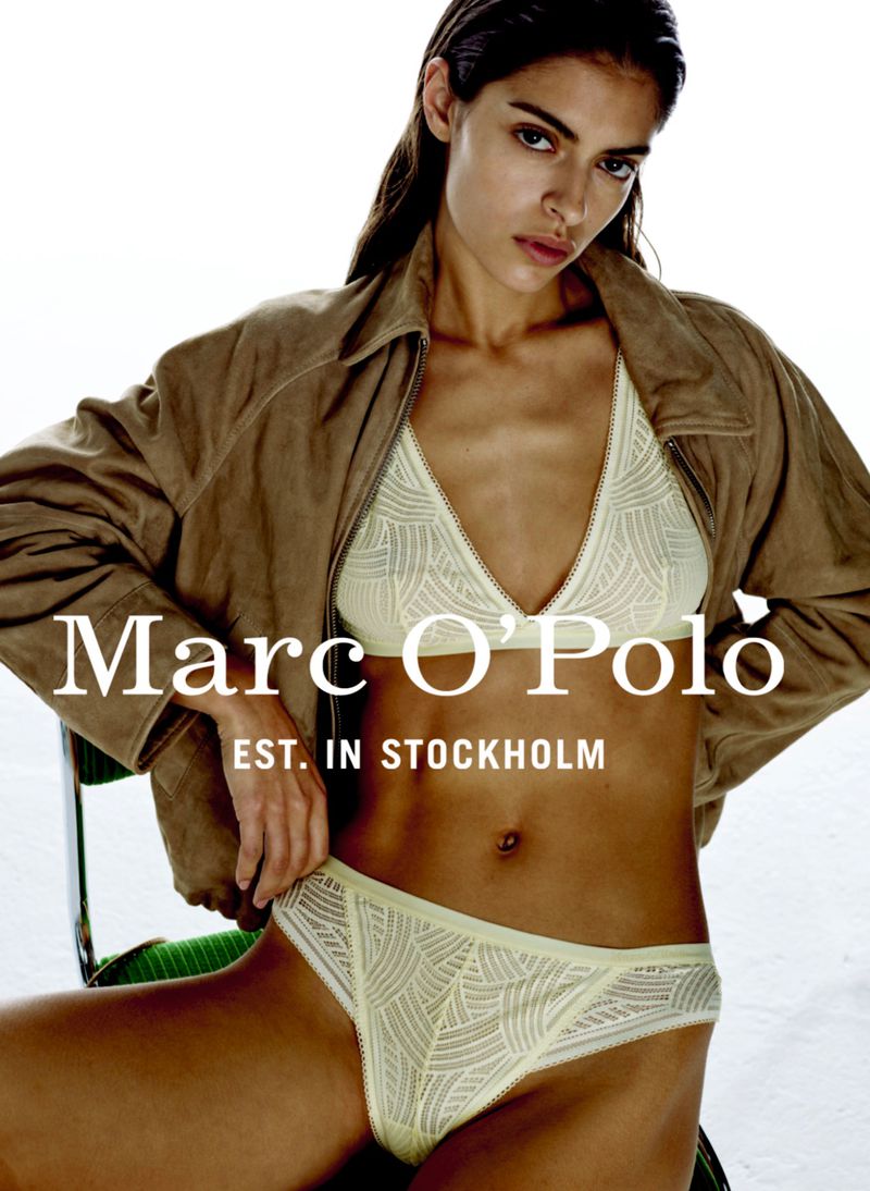 A woman poses wearing a beige jacket and ivory lingerie set. The Marc O’Polo logo is displayed on the left side of the image.