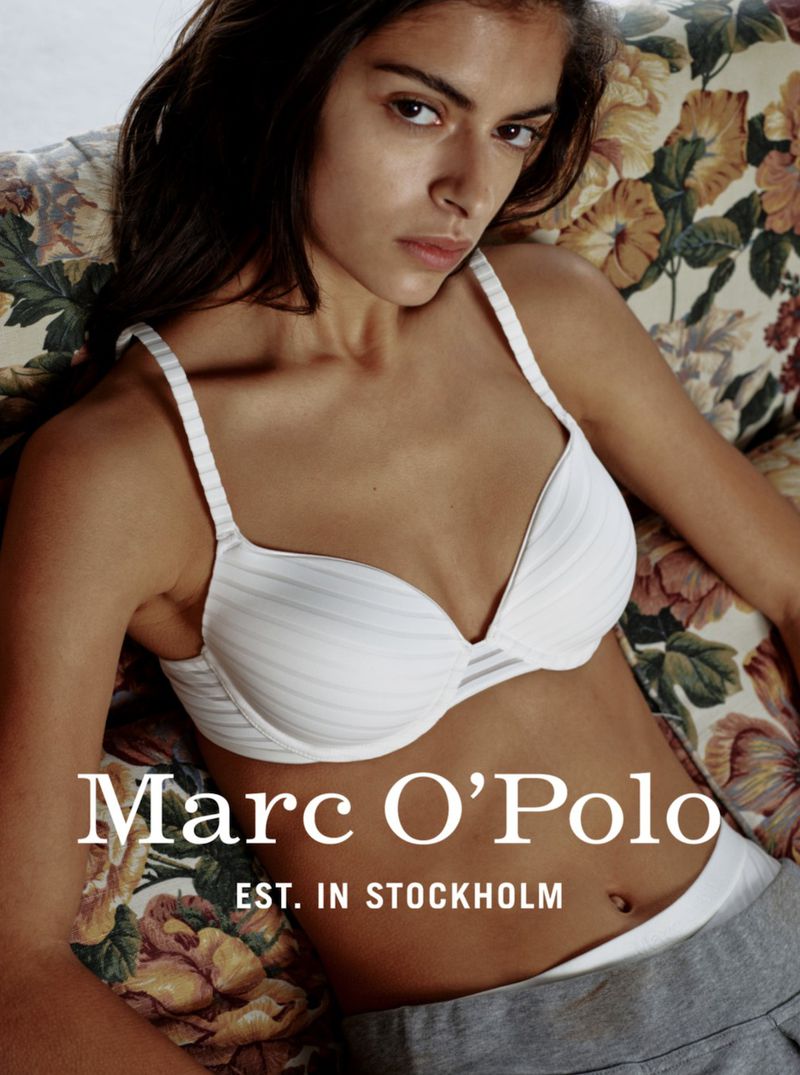 Indoor portrait of a woman in a bra and grey pants, lounging on a floral patterned couch with the 'Marc O' Polo' logo overlaid on the image.