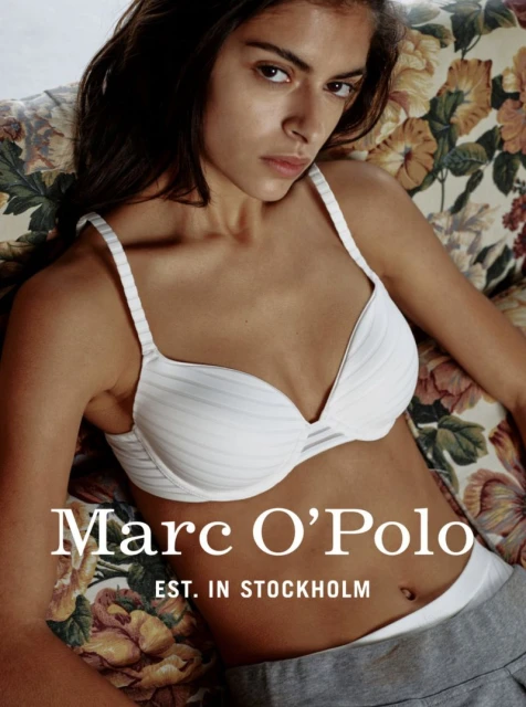 Indoor portrait of a woman in a bra and grey pants, lounging on a floral patterned couch with the 'Marc O' Polo' logo overlaid on the image.