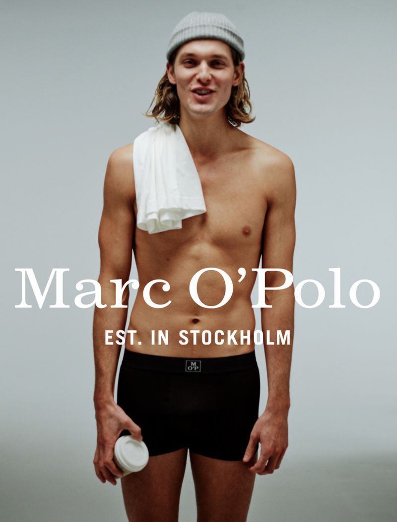Young man in underwear and a beanie poses in a studio, holding a coffee cup, with the Marc O'Polo logo prominently displayed.