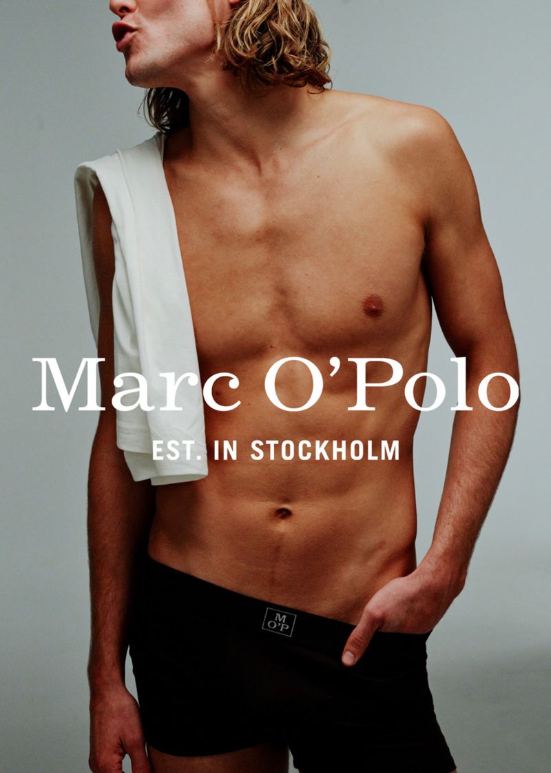 Male model posing shirtless with black underwear and a white towel, set against a grey background in a fashion advertisement.