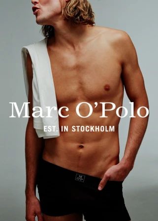 Male model posing shirtless with black underwear and a white towel, set against a grey background in a fashion advertisement.